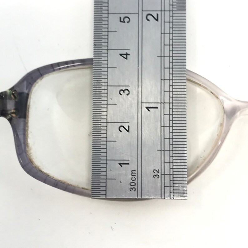 May include: Close-up of a pair of eyeglasses with a ruler measuring the lens. The lens is approximately 1 inch wide. The frame is a translucent gray color.