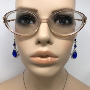 May include: Oval-shaped eyeglasses with a light brown frame and gold accents. The glasses are on a mannequin head, paired with dangling blue teardrop earrings. The mannequin has full lips and long eyelashes.