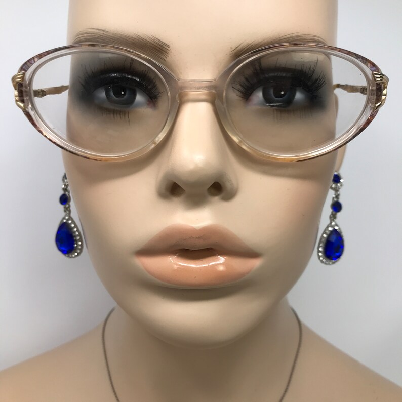 May include: Oval-shaped eyeglasses with a light brown frame and gold accents. The glasses are on a mannequin head, paired with dangling blue teardrop earrings. The mannequin has full lips and long eyelashes.