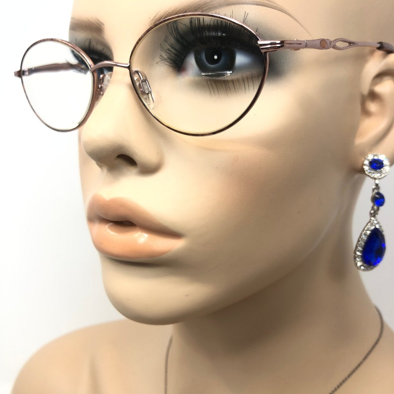 K&ouml;nnte beinhalten: Ros&eacute;goldene ovale Brille mit klaren Gl&auml;sern. Die Brille befindet sich auf einer Schaufensterpuppe. Ein Paar blaue und silberne Ohrh&auml;nger sind rechts zu sehen. Die Ohrringe haben ein Blumenmuster und einen gro&szlig;en tropfenf&ouml;rmigen blauen Stein.