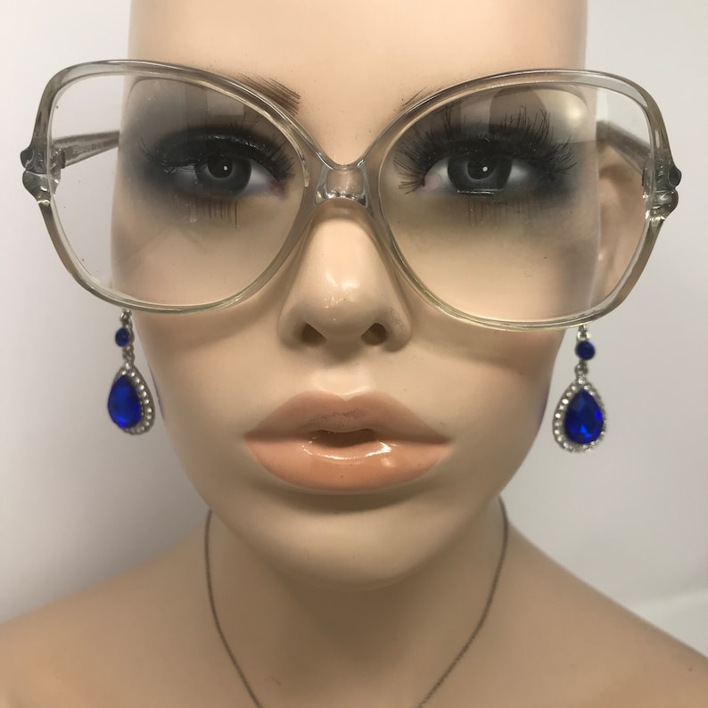 May include: Clear oversized eyeglasses with a light frame. The glasses are displayed on a mannequin with long eyelashes and a pair of blue teardrop earrings with silver accents.