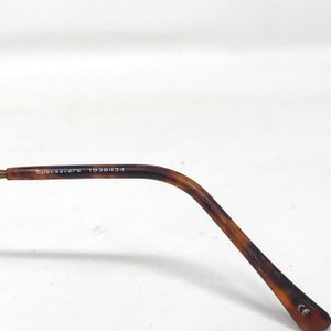 May include: Close-up of a brown and tan tortoiseshell eyeglasses arm. The text "Specsavers 1938434" is printed on the arm. The arm is curved and has a rounded tip.
