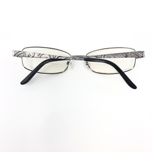 May include: Silver-framed eyeglasses with a decorative pattern on the temples and black arms. The lenses are clear, and the glasses are set against a white background.