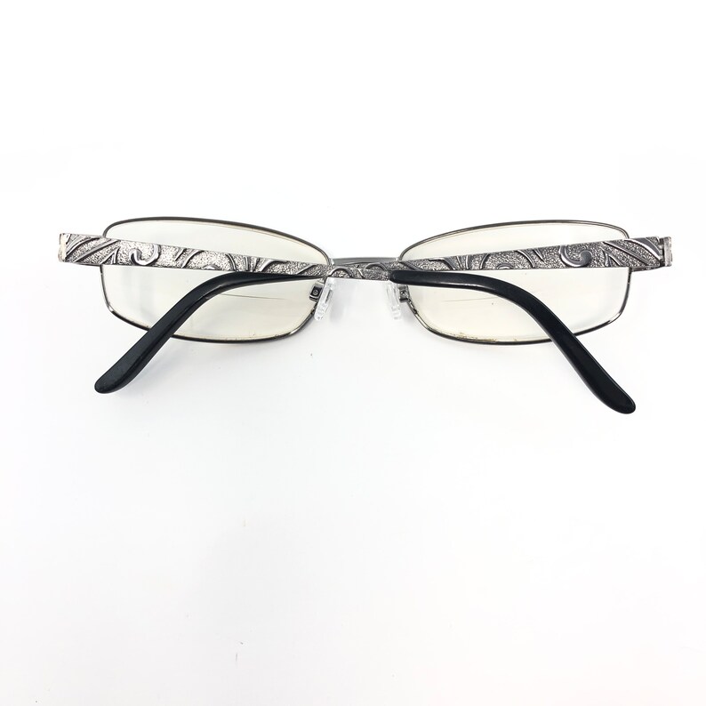 May include: Silver-framed eyeglasses with a decorative pattern on the temples and black arms. The lenses are clear, and the glasses are set against a white background.