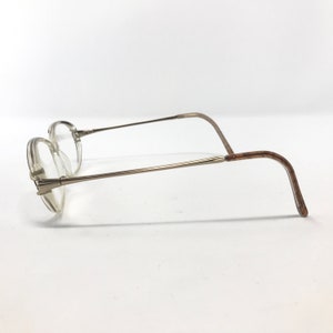 K&ouml;nnte beinhalten: Eine Brille mit einem d&uuml;nnen, silberfarbenen Metallrahmen. Die Gl&auml;ser sind klar, und die B&uuml;gel biegen sich, um hinter den Ohren zu liegen. Die Brille ist vor einem wei&szlig;en Hintergrund platziert.