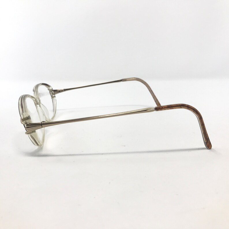 K&ouml;nnte beinhalten: Eine Brille mit einem d&uuml;nnen, silberfarbenen Metallrahmen. Die Gl&auml;ser sind klar, und die B&uuml;gel biegen sich, um hinter den Ohren zu liegen. Die Brille ist vor einem wei&szlig;en Hintergrund platziert.