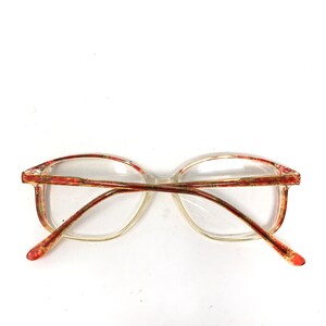 May include: Clear eyeglasses with a rectangular frame and red and gold accents. The arms of the glasses are a reddish-brown color. The lenses are clear.