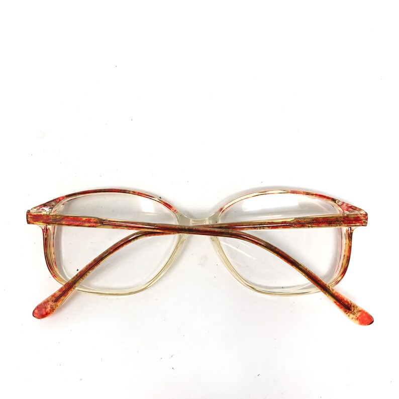 May include: Clear eyeglasses with a rectangular frame and red and gold accents. The arms of the glasses are a reddish-brown color. The lenses are clear.