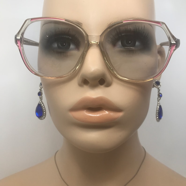 May include: Clear eyeglasses with a pink and gold frame. The glasses have a large, octagonal shape. The mannequin is also wearing silver earrings with blue teardrop-shaped gems. The mannequin has a neutral skin tone and is set against a white background.