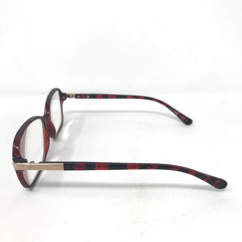 May include: A pair of eyeglasses with a tortoiseshell pattern on the frames and temples. The frames are a reddish-brown color, and the temples have a black and red striped design. The glasses have a gold-colored metal accent at the hinges.