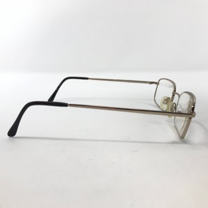 May include: Silver-framed eyeglasses with rectangular lenses. The glasses have thin silver arms and black earpieces. The lenses are clear, and the nose pads are a light beige color. The glasses are set against a white background.
