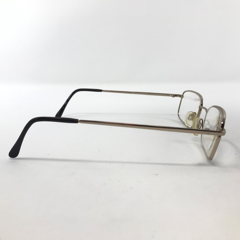 May include: Silver-framed eyeglasses with rectangular lenses. The glasses have thin silver arms and black earpieces. The lenses are clear, and the nose pads are a light beige color. The glasses are set against a white background.
