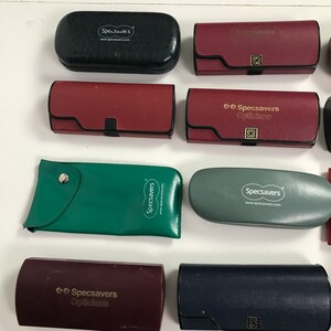 May include: Assortment of Specsavers eyeglass cases in various colors, including black, burgundy, green, and gray. The cases are rectangular and oval-shaped, with the Specsavers logo printed on them. Some cases have a snap closure.