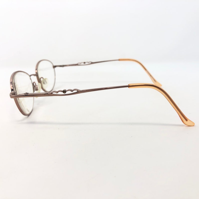 May include: A pair of eyeglasses with a brown metal frame and oval lenses. The temples are thin and extend to a gold-colored ear piece. The frame features a decorative heart-shaped design near the hinges.
