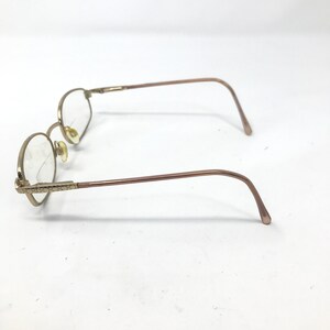 May include: Gold-coloured, round-framed spectacles with clear lenses. The frames have a delicate, vintage aesthetic, with decorative detailing on the temples. The arms curve gently, and the nose pads are a light beige colour.