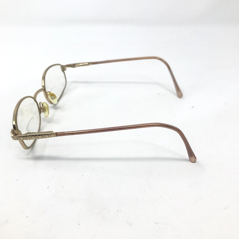 May include: Gold-coloured, round-framed spectacles with clear lenses. The frames have a delicate, vintage aesthetic, with decorative detailing on the temples. The arms curve gently, and the nose pads are a light beige colour.