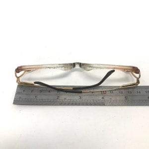 May include: Clear eyeglasses with a gold-colored frame and black earpieces. The glasses are positioned next to a ruler, with a measurement of approximately 5 inches.