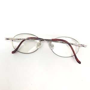 May include: A pair of oval-shaped spectacles with a silver metal frame and brown plastic arms. The frame has a decorative design with a purple and gold pattern.