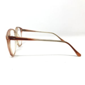 May include: A pair of eyeglasses with a light brown frame and clear lenses. The glasses have a rectangular shape and a thin, metal arm that transitions from silver to brown. The glasses are set against a white background.