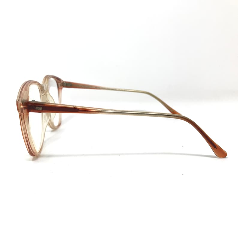 May include: A pair of eyeglasses with a light brown frame and clear lenses. The glasses have a rectangular shape and a thin, metal arm that transitions from silver to brown. The glasses are set against a white background.