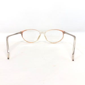 May include: Oval-framed spectacles with a transparent frame and peach-coloured gradient accents. The arms are silver-toned. The lenses are clear. The glasses are set against a white background.