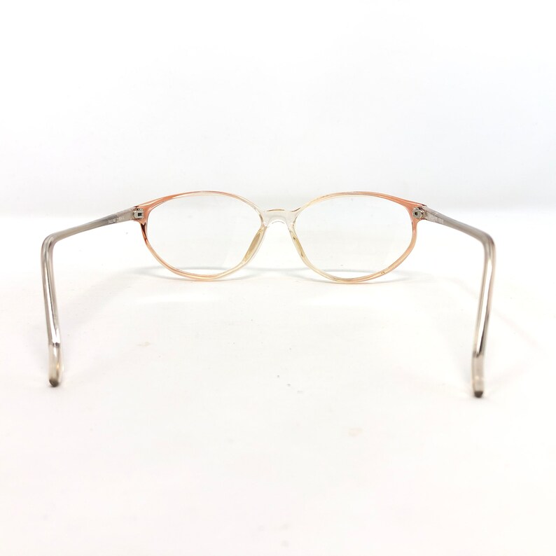 May include: Oval-framed spectacles with a transparent frame and peach-coloured gradient accents. The arms are silver-toned. The lenses are clear. The glasses are set against a white background.