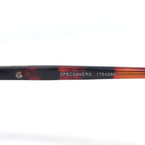 May include: Close-up of a portion of eyeglasses with a tortoiseshell pattern. The text "SPECSAVERS 1764286" is printed on the arm of the glasses. The frame is brown and orange.