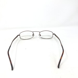 May include: Brown metal eyeglasses with rectangular lenses and a thin frame. The temples are a darker brown with a tortoise shell pattern. The glasses are set against a white background.