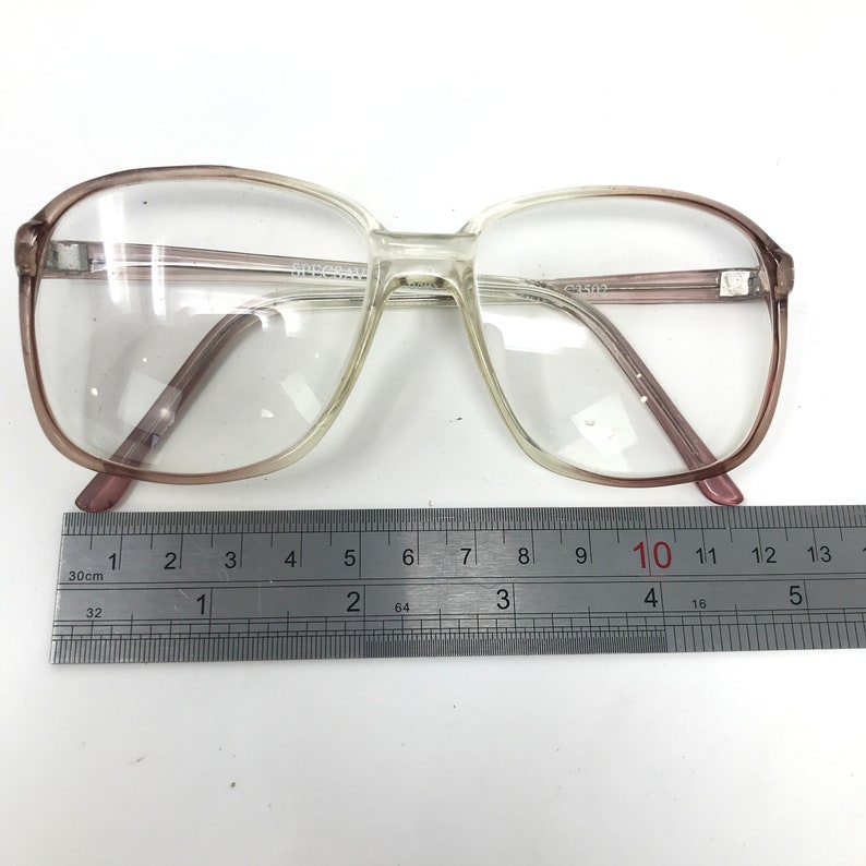 May include: Clear eyeglasses with a light brown gradient frame. The glasses have a rectangular shape and are placed on a white surface next to a ruler. The frame measures approximately 5 inches in width.