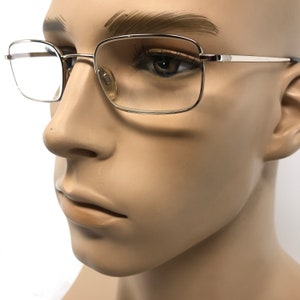May include: Silver-framed eyeglasses with a rectangular lens shape. The glasses have a thin, metallic frame and clear lenses. The nose pads are a light beige color. The glasses are on a mannequin head.