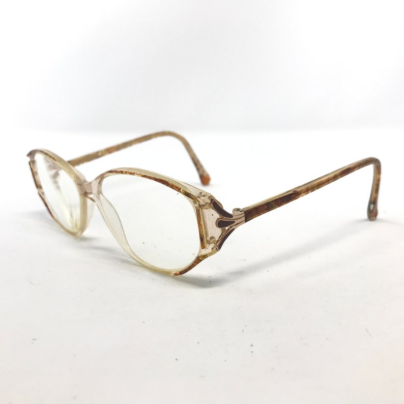 May include: Vintage eyeglasses with a clear, rectangular frame and brown temples. The lenses are clear, and the frame has a slight pinkish tint. The glasses have a classic, retro design.