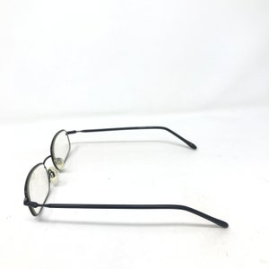 Puede incluir: Gafas con montura de metal negro y lentes ovaladas. Las gafas tienen una montura y patillas finas, y almohadillas nasales. Las lentes son transparentes. Las gafas est&aacute;n sobre un fondo blanco.