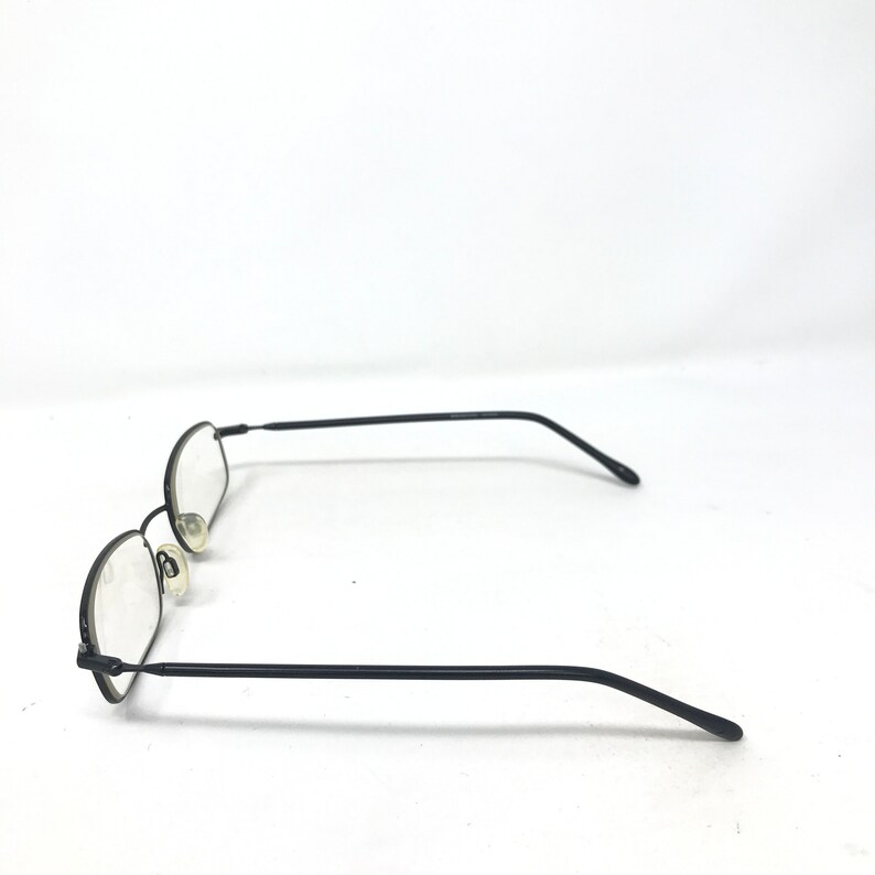 Puede incluir: Gafas con montura de metal negro y lentes ovaladas. Las gafas tienen una montura y patillas finas, y almohadillas nasales. Las lentes son transparentes. Las gafas est&aacute;n sobre un fondo blanco.