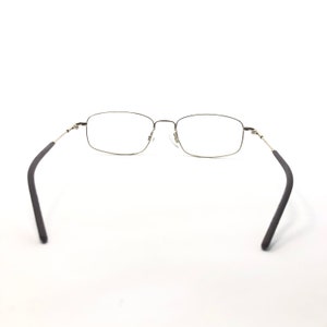 May include: A pair of rectangular eyeglasses with a silver metal frame and black temple arms. The lenses are clear, and the glasses are set against a white background. The glasses are designed for vision correction.