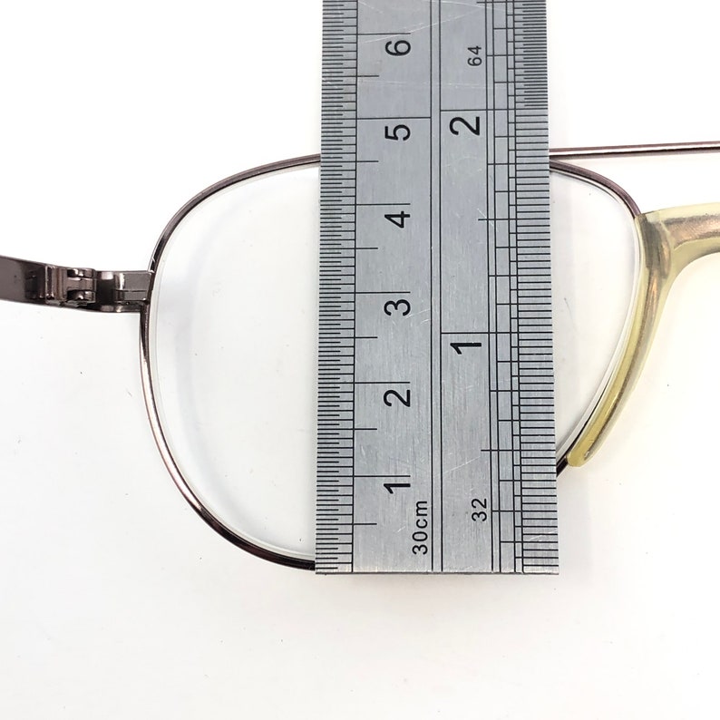 May include: Close-up of eyeglasses with a silver metal frame. A ruler is placed next to the lens, showing a measurement of approximately 1.5 inches. The glasses have a clear lens and a light-colored arm.