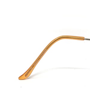 Puede incluir: Primer plano de una patilla de gafas dorada. El brazo se curva suavemente y tiene texto impreso. El texto incluye los n&uacute;meros "24336944" y "110-13K". El brazo est&aacute; sobre un fondo blanco.
