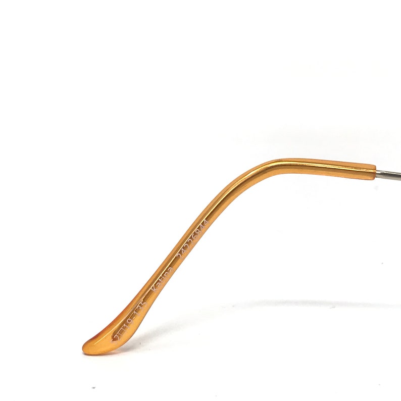 Puede incluir: Primer plano de una patilla de gafas dorada. El brazo se curva suavemente y tiene texto impreso. El texto incluye los n&uacute;meros "24336944" y "110-13K". El brazo est&aacute; sobre un fondo blanco.