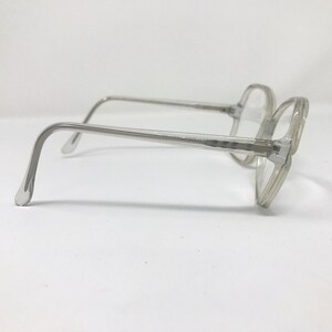 May include: Clear, square-framed eyeglasses with a light gray tint. The glasses have a classic design with a thick frame and temples. The lenses are clear, and the overall look is vintage.