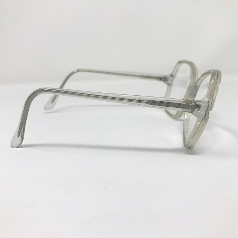 May include: Clear, square-framed eyeglasses with a light gray tint. The glasses have a classic design with a thick frame and temples. The lenses are clear, and the overall look is vintage.