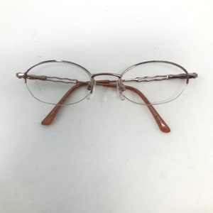May include: A pair of oval-shaped eyeglasses with a thin, metallic frame in a light brown hue. The lenses are clear, and the temples are a darker brown. The glasses have a minimalist design, suitable for everyday wear.