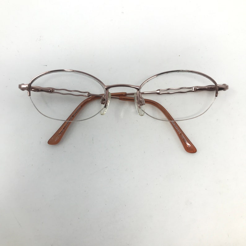 May include: A pair of oval-shaped eyeglasses with a thin, metallic frame in a light brown hue. The lenses are clear, and the temples are a darker brown. The glasses have a minimalist design, suitable for everyday wear.