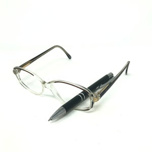 May include: Clear-framed spectacles and a black pen. The spectacles have a cat-eye shape with a light brown gradient on the arms. The pen is black with silver detailing. The items are on a white background.