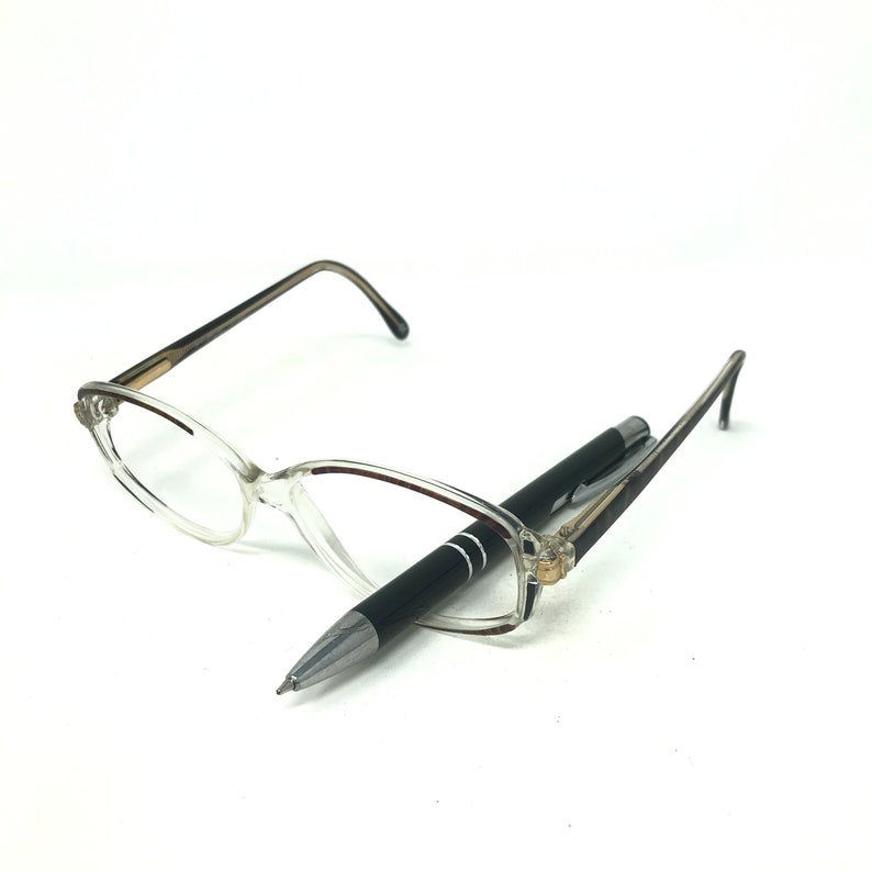 May include: Clear-framed spectacles and a black pen. The spectacles have a cat-eye shape with a light brown gradient on the arms. The pen is black with silver detailing. The items are on a white background.