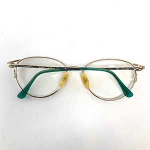 Pu&ograve; includere: Occhiali in metallo dorato con aste in plastica verde e un motivo floreale sulla montatura.