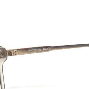 May include: Close-up of a pair of eyeglasses with a clear frame and gold-colored arms. The text "SPECSAVERS 1044267" is printed on the arm of the glasses. The glasses are set against a white background.