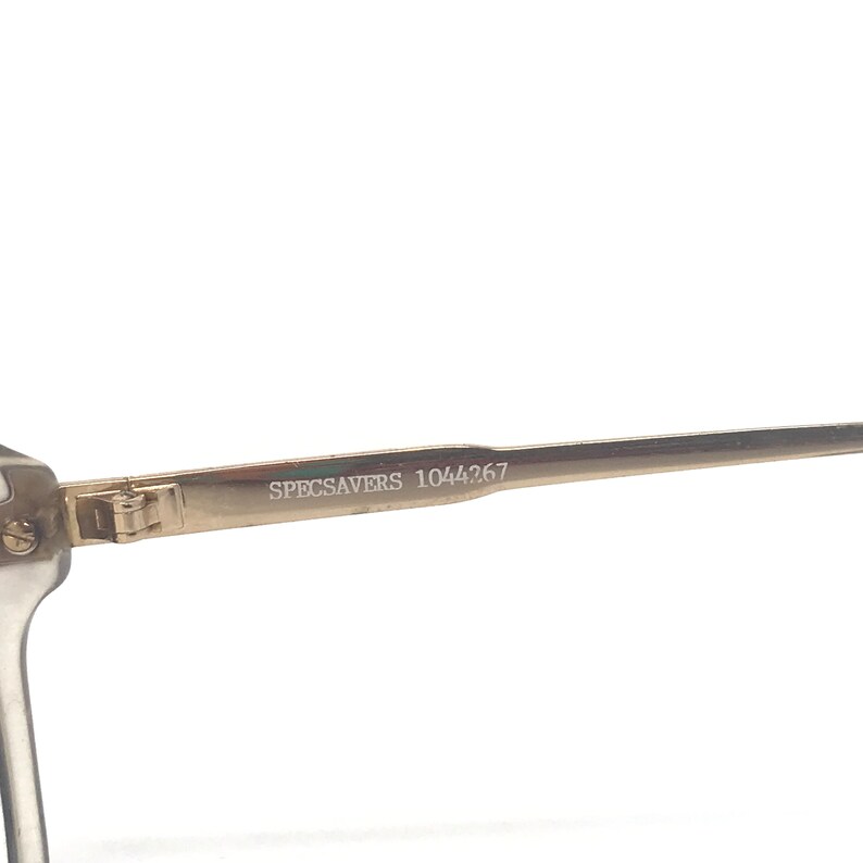 May include: Close-up of a pair of eyeglasses with a clear frame and gold-colored arms. The text "SPECSAVERS 1044267" is printed on the arm of the glasses. The glasses are set against a white background.