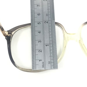 May include: A pair of eyeglasses with a silver frame, measured with a ruler. The ruler shows the frame is approximately 2 inches wide. The lenses are clear, and the arms of the glasses are a light color.