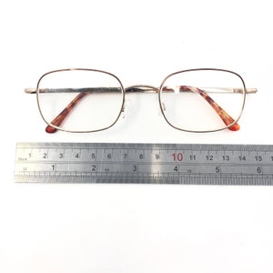 May include: Rectangular eyeglasses with clear lenses and a gold-coloured frame. The arms are brown tortoiseshell. A metal ruler is placed below the glasses, with measurements in centimetres.