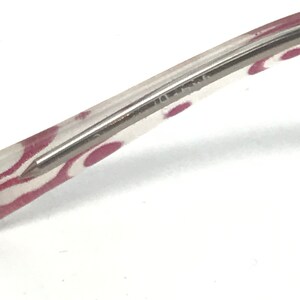 May include: Close-up of a clear eyeglass frame with a silver metal arm. The frame has a pattern of pink ovals. The arm is thin and rectangular, with a pointed tip.