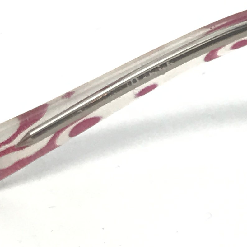 May include: Close-up of a clear eyeglass frame with a silver metal arm. The frame has a pattern of pink ovals. The arm is thin and rectangular, with a pointed tip.